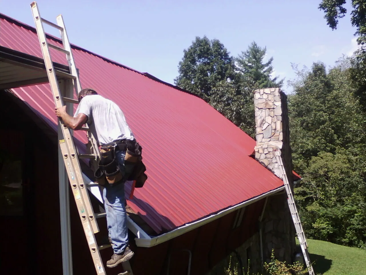 Roofer on a ladder beside a red metal roof and stone chimney — Soffit & Fascia Repair by Roofing Brothers in Shawmut